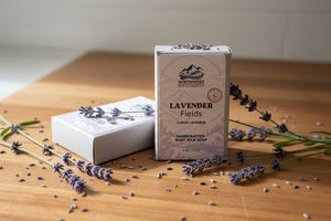 A rectangular bar of Northwest Soap Co. Lavender Fields Classic Lavender goat milk soap, shown in its paper packaging with a second soap bar partially visible behind it on a woood counter with lavender sprigs.
