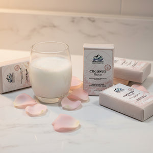 A rectangular bar of Northwest Soap & Candle Co. Coconut Rose handcrafted coconut milk soap, shown in its kraft paper packaging with a white and pink label, set on a counter with a glass of milk and rose petals.