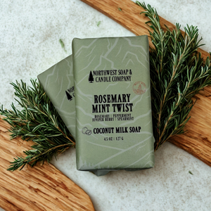 Two bars of Northwest Soap & Candle Company Rosemary Mint Twist soap with rosemary leaves on a wooden surface.